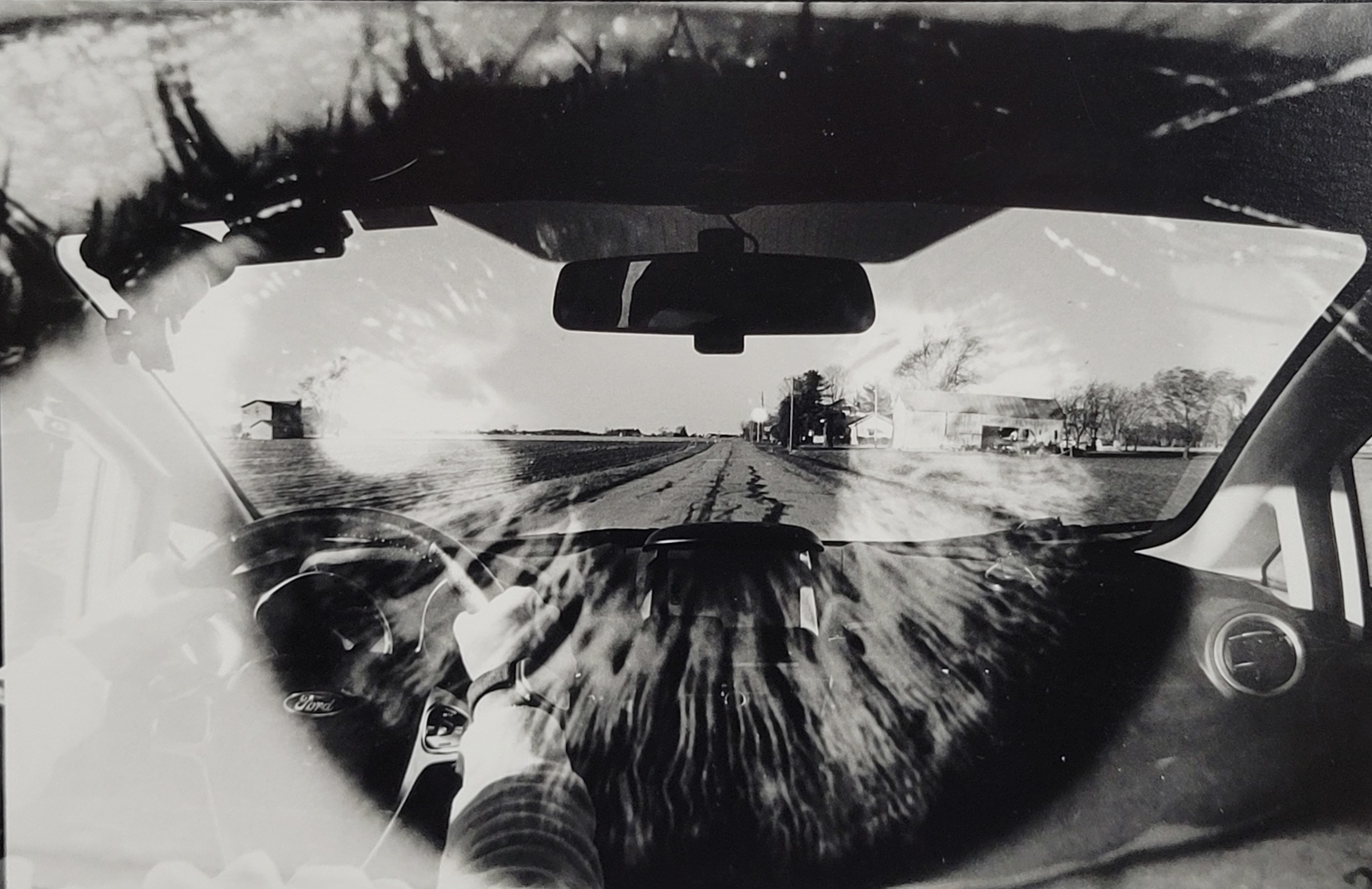 A black and white photograph. The perspective is inside a car looking out the front windshield, the driver on the left side of the image. A country road with wide farmland is outside the car, with the horizon line centered in the photo. A second image is overlayed over with the image of an eye, the end of the road to the horizon is centered in the iris of the eye.