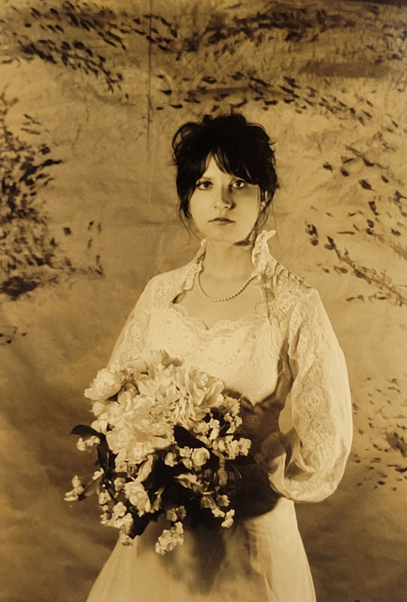 A sepia toned photograph of a woman in Victorian era clothing. A white, laced dress with a high collar faces the camera. A bouquet of flowers in her hand, making her appear bride-like. Floral elements are behind her in the backdrop