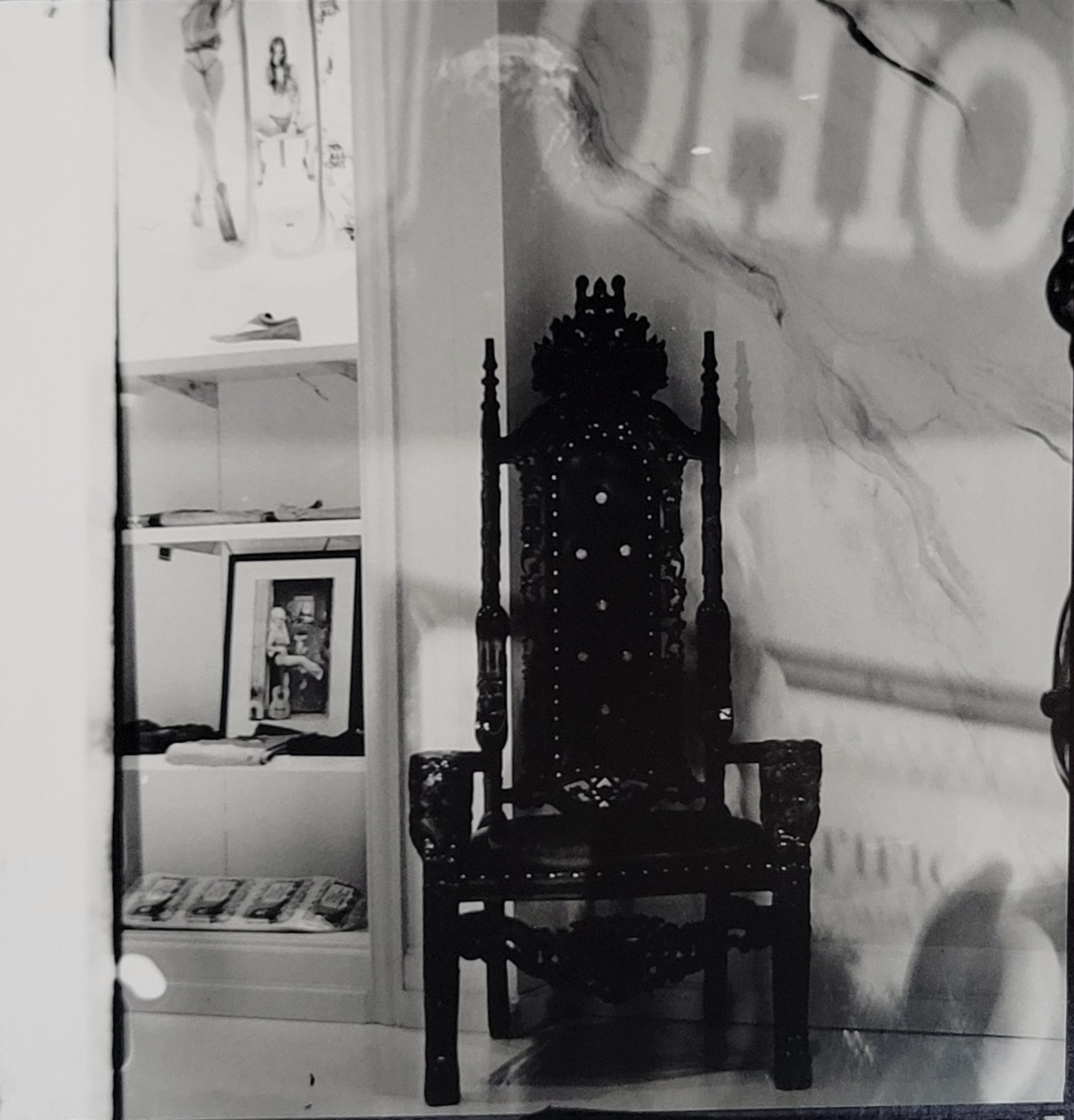 A black and white photograph of a large, ornate black chair sitting in a white room. A tall back with lots of ornamentation make the chair look like a throne.