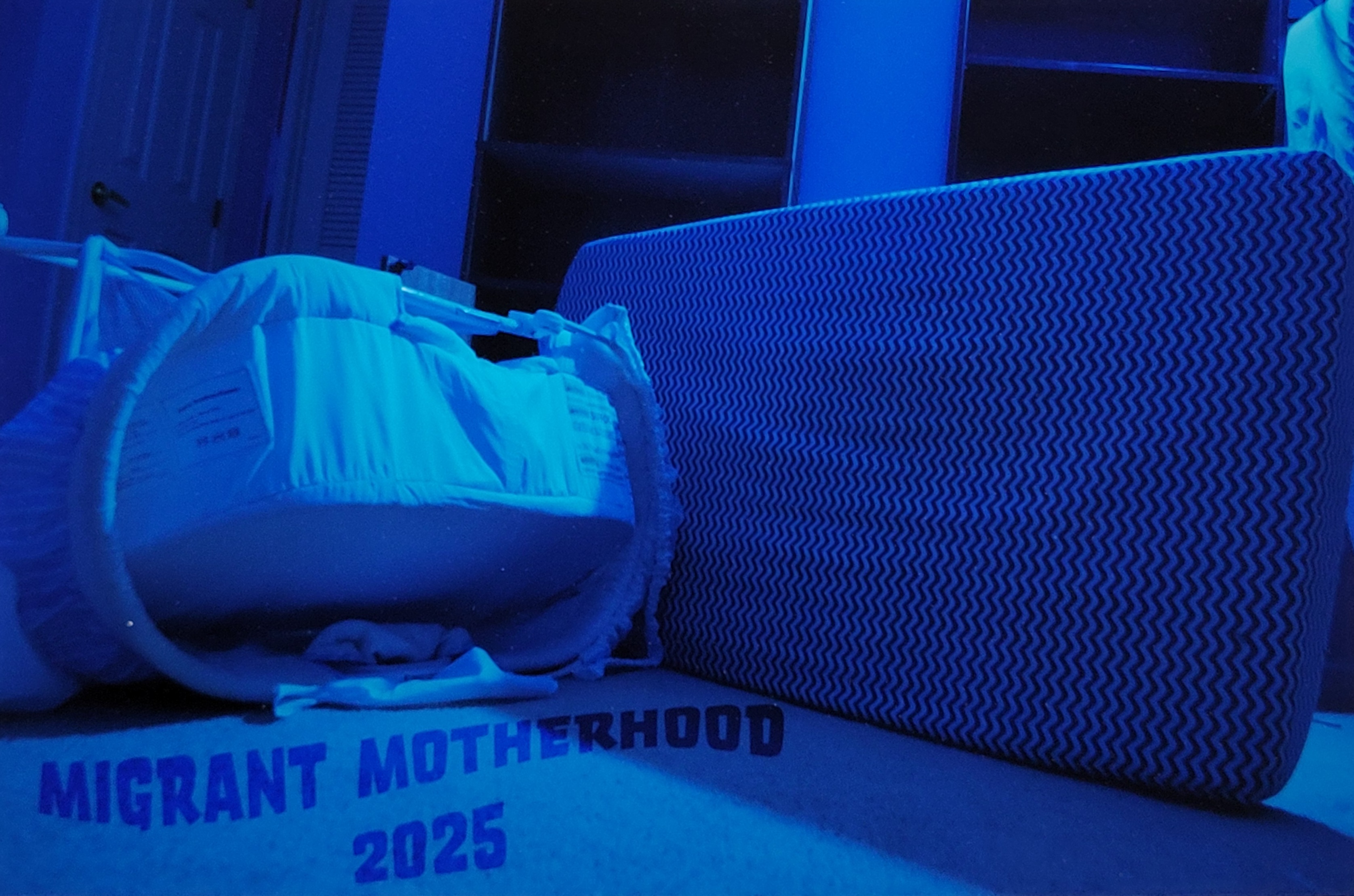 A digital color photograph tinted blue. It depicts a mattress and a baby crib thrown on their sides. The words "Migrant Motherhood 2025" are digitally placed in the lower left corner beneath the crib.