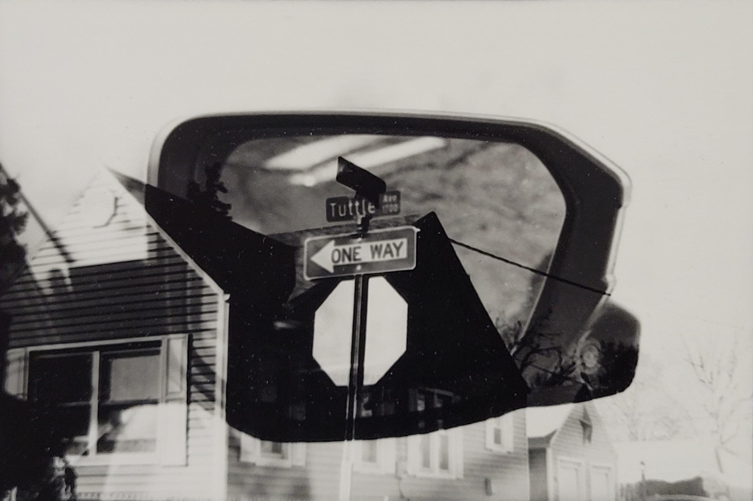 Black and white photo of a side view mirror with a stop sign juxtaposed over the mirror, making it appear that it is in the side mirror.