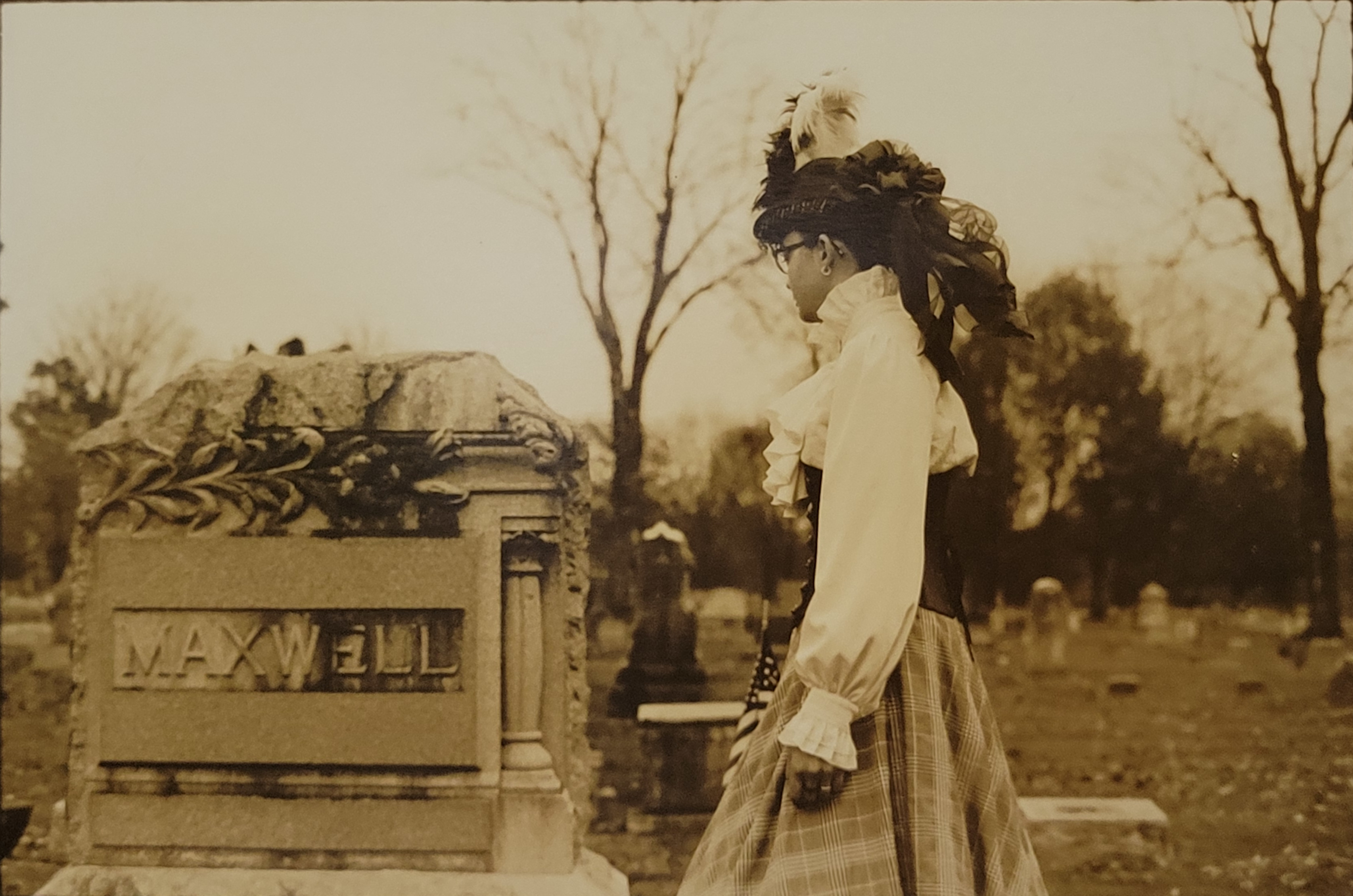 A sepia toned photograph depicting a young woman in period attire, around 1870, looking away from the camera at a headstone. She stands in a cemetery with trees and other headstones behind her.