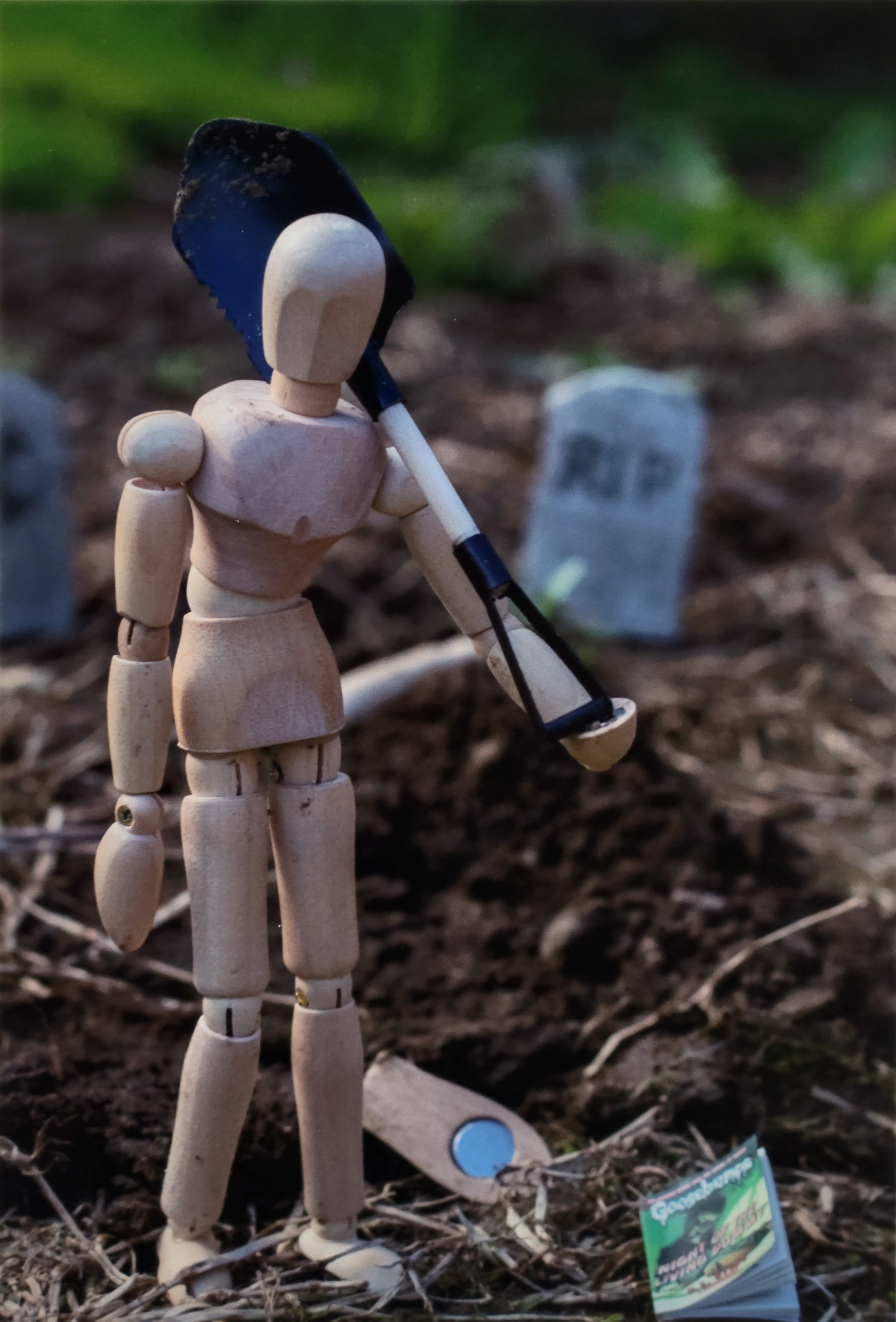 A color photograph of a still life scene. A wooden model figure stands in the foreground of a cemetary, holding a shovel with it leaning on its shoulder. A Goosebumps book titled "Night of the Living Dummy" beside the figure. Directly behind the wooden figure is a freshly buried wooden figure with only its foot sticking out.