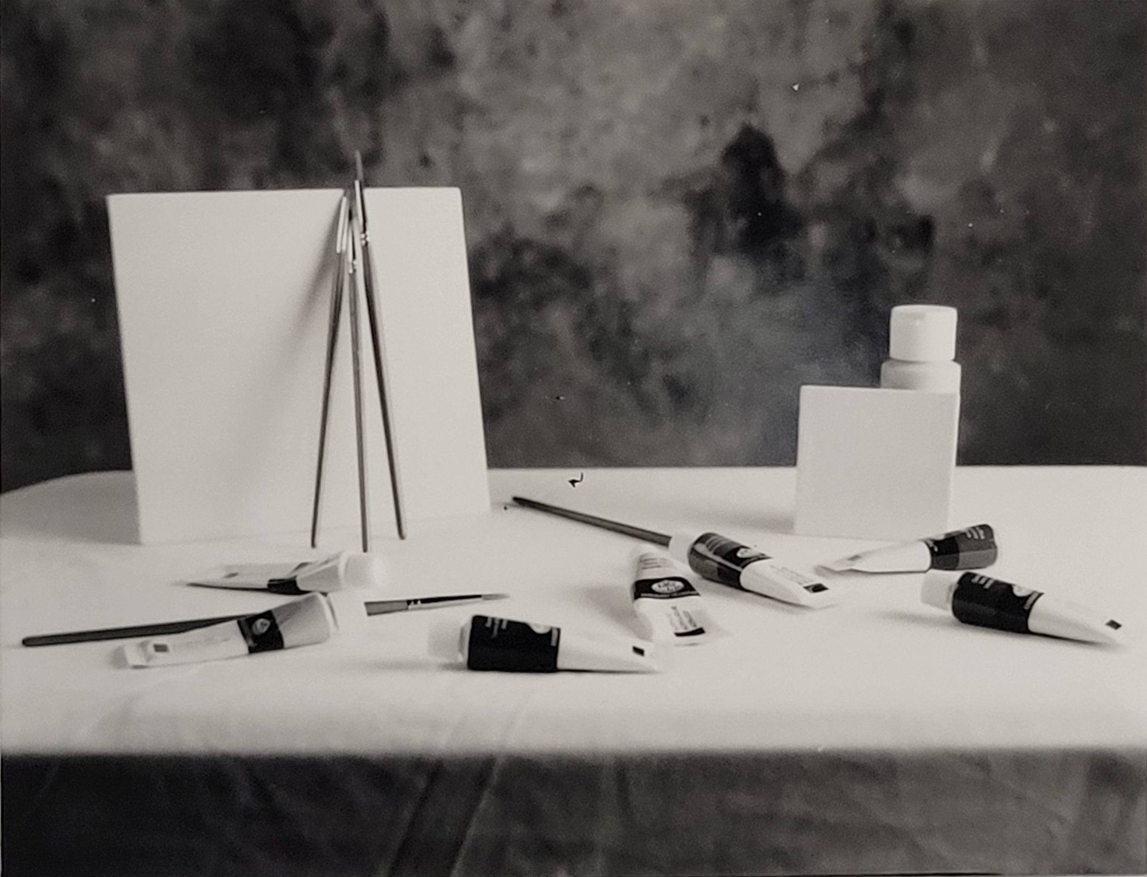 A still life black and white photo. A white covered table, two blank canvases are propped up on either side of the photo. Paintbrushes lean against the canvas on the left. In front of the canvases are paint tubes strewn across the table.