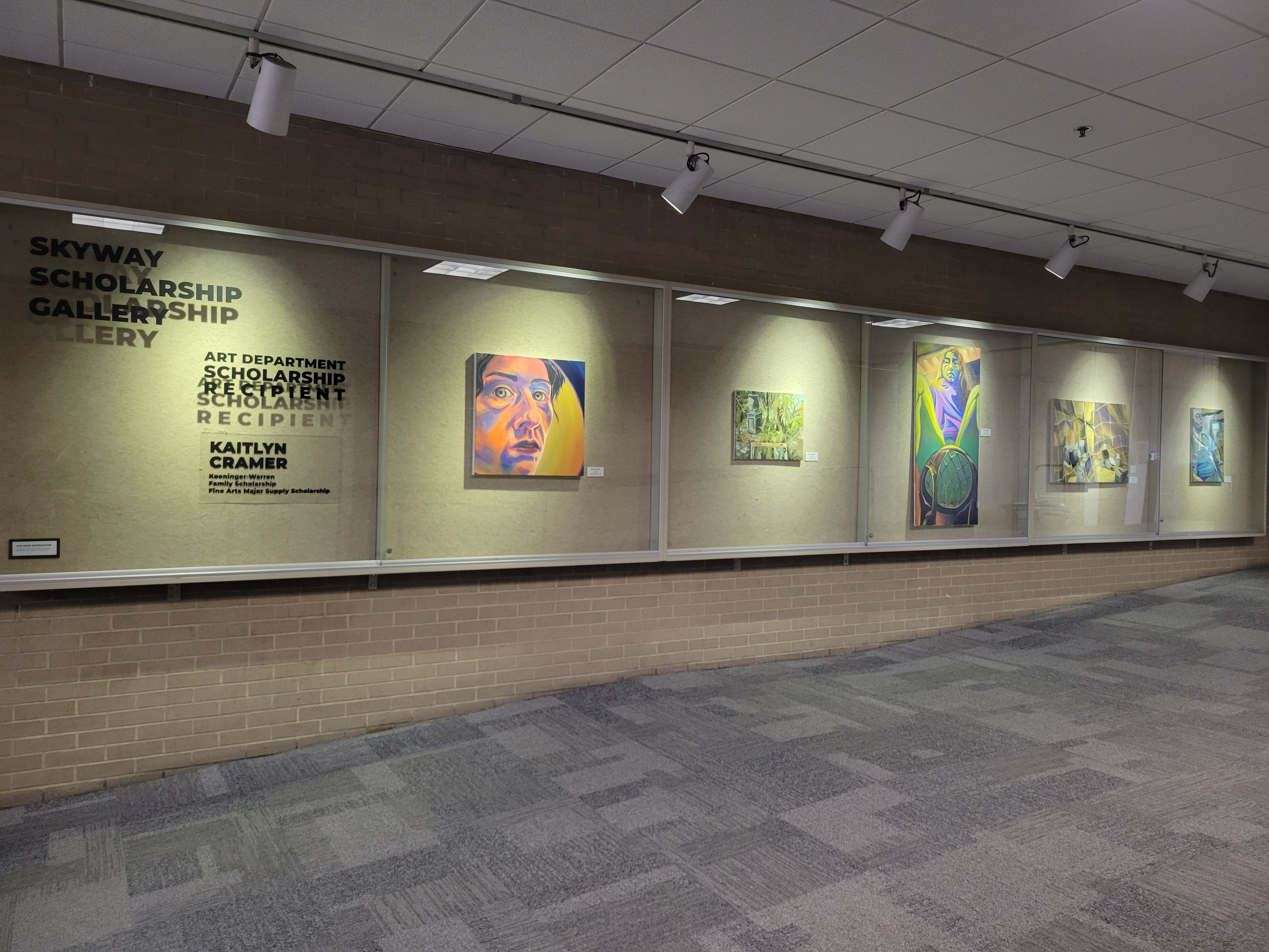 Skyway Gallery displaying work by student artist. It is a thin gallery along the wall, with 5 pieces in each section, the 6th section is the title of the gallery and the featured students name.