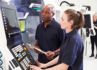 Apprenticeship Skill Training photo is depicting a mentor with a clipborad in hand, standing and watching and carefully monitoring the various steps the girl apprentice standing next to him is performing on an industial programing console