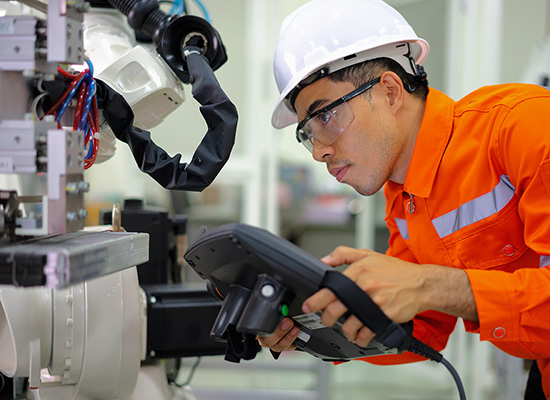SMART Manufacturing Skill Training image is depicting a young man apprentice in an orange color (industrial used) outfit, with a white color hard hard while leaning over some industrial equipment to read data that he is inputing on handheld device on hand
