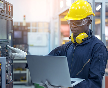 Information for prospective students is depicting an apprentice in training wearing a yellow hard hat, he is holding a laptop in left arm while punching in on the keypad of an automation control unit with his right hand and viewing values to input from his laptop.
