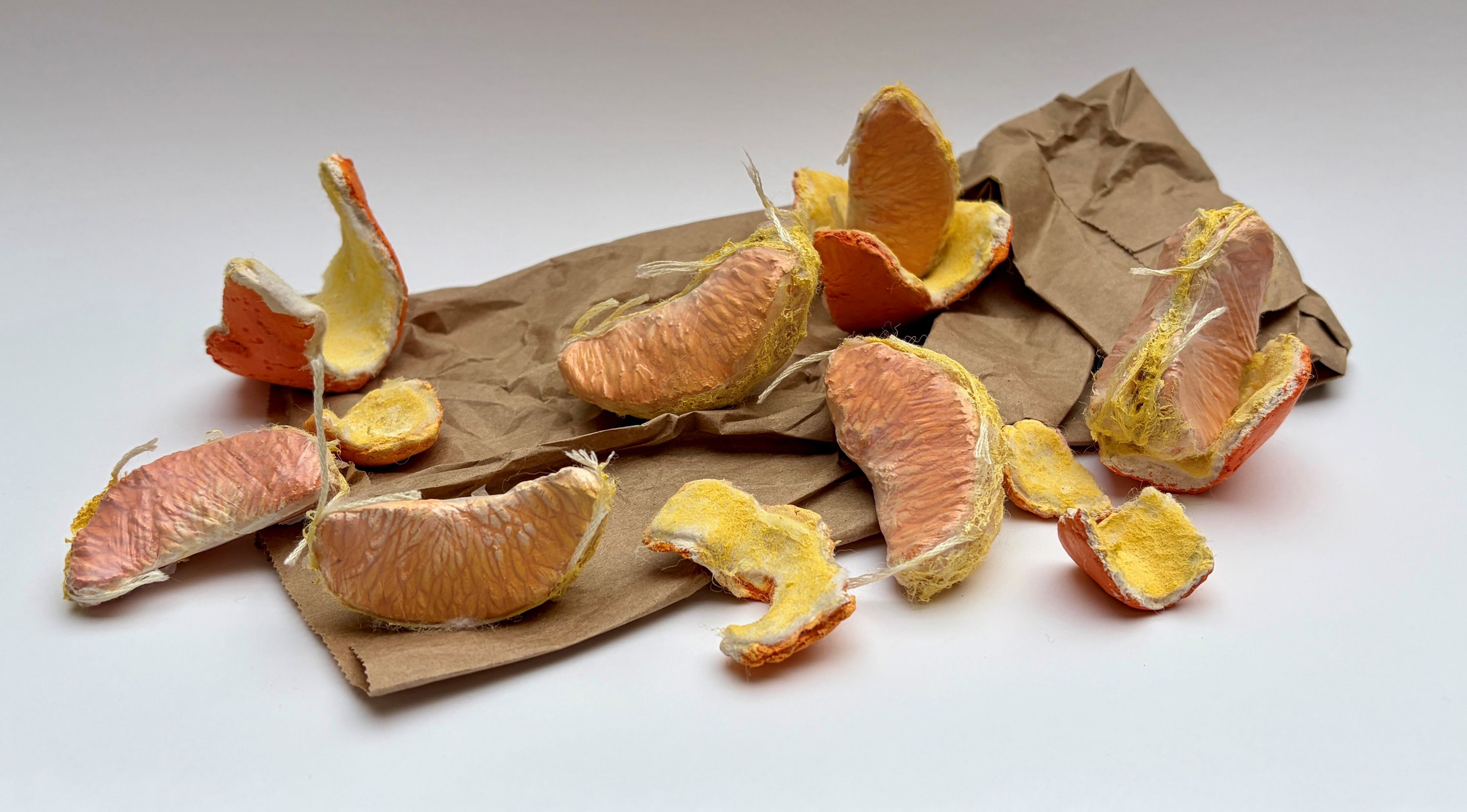 A sculptural replica of a peeled orange. The orange slices and the orange skin pieces sit on top of a paper bag.