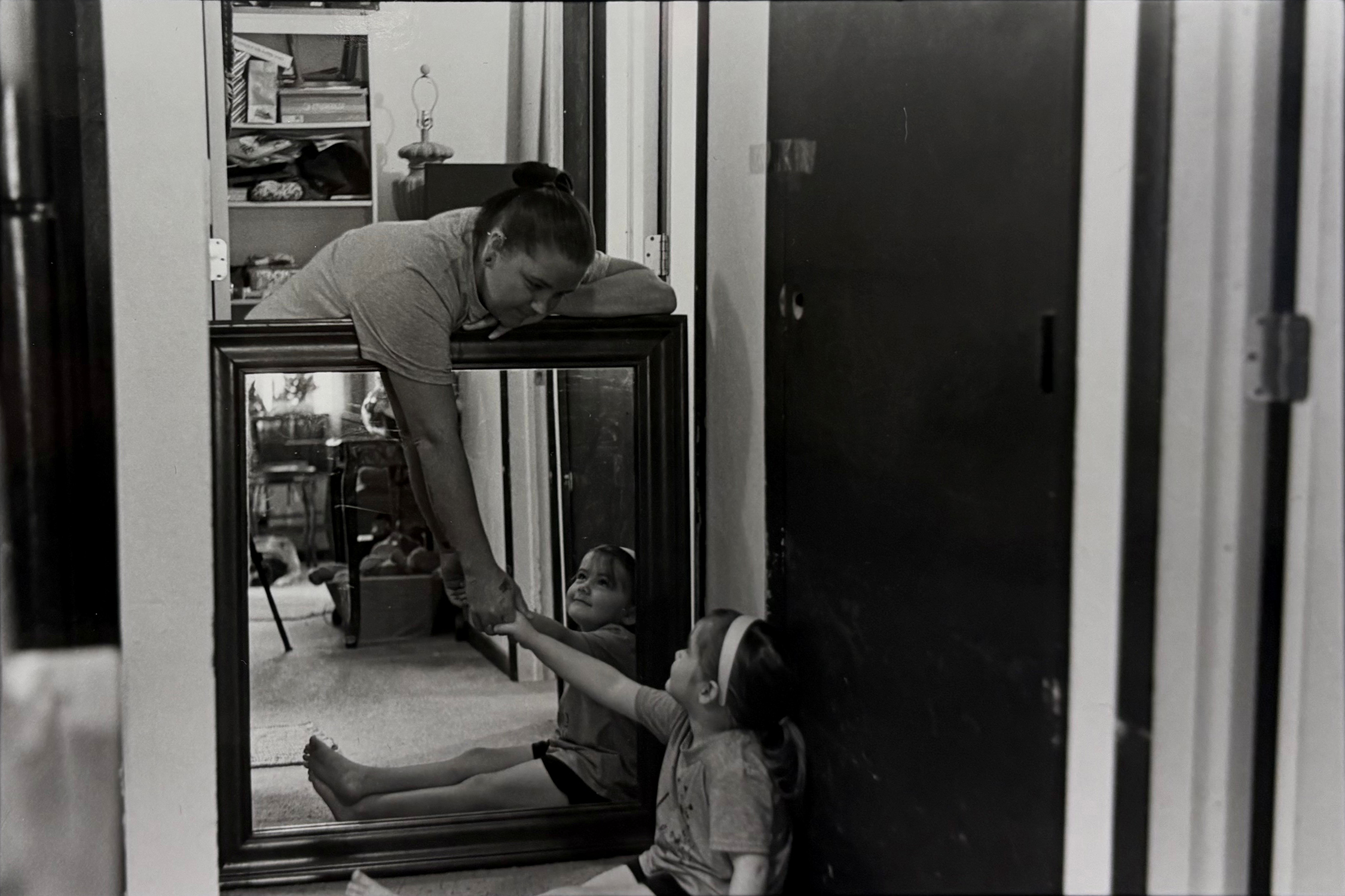 A black and white photograph of a mother leaning down over a mirror to hold the hand of her young daughter who is sitting on the ground. The mirror reflects the daughter, who is fondly looking up to her mother. Set in the interior of a home, there are many verticals of door jambs contrasting to the diagonal of the arms and forms of the mother and daughter.