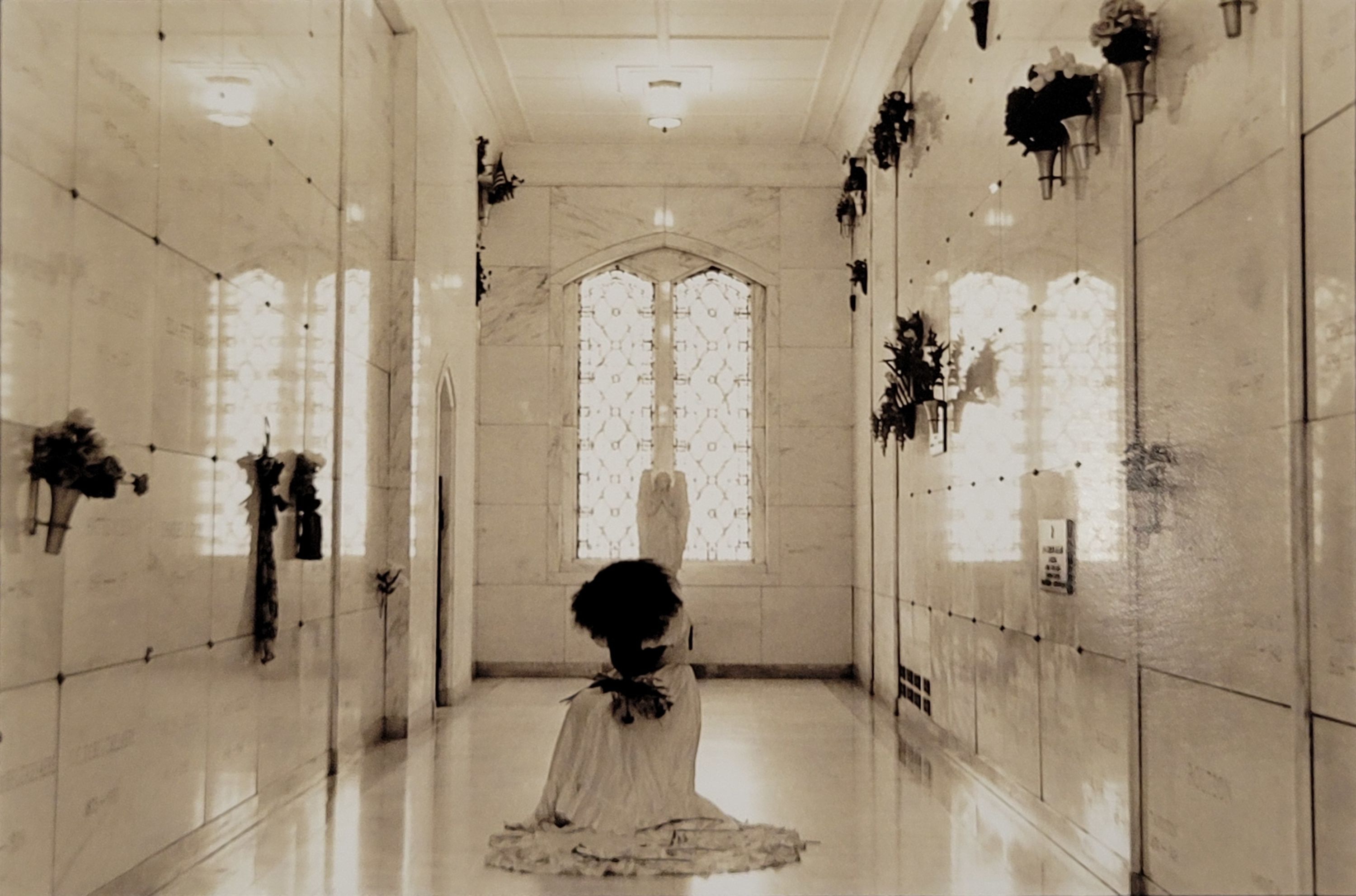 A sepia toned photograph depicting a woman in a wedding dress, hunched over on the floor holding herself. She is centered inside a mausoleum, marble graves and flowers on either side of her. Her thick hair covers her face as she droops her head downward. Behind her are ornate windows on either side of a marble statue of an angel, giving the appearance that the angel is watching over the woman. 