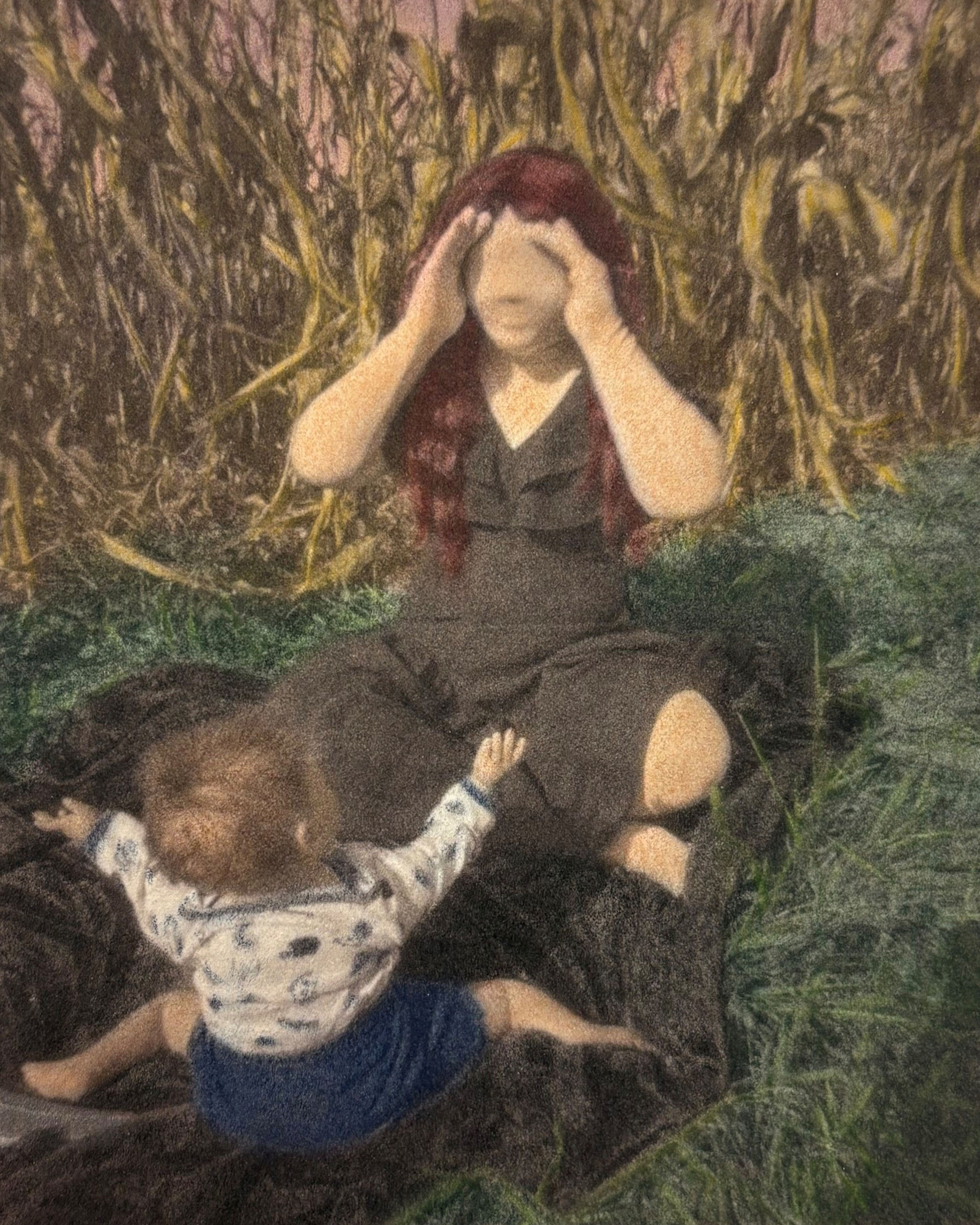 A faceless female figure sits in a corn field in an open patch. She sits on a black blanket, playing peek-a-boo with a small child. The child has their arms outstretched in excitement.