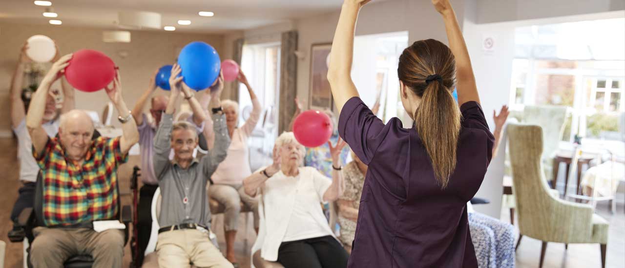 Senior facility members sit in chairs while doing exercises involving holding up balloons over their heads. A staff member demonstrates the exercise to them.